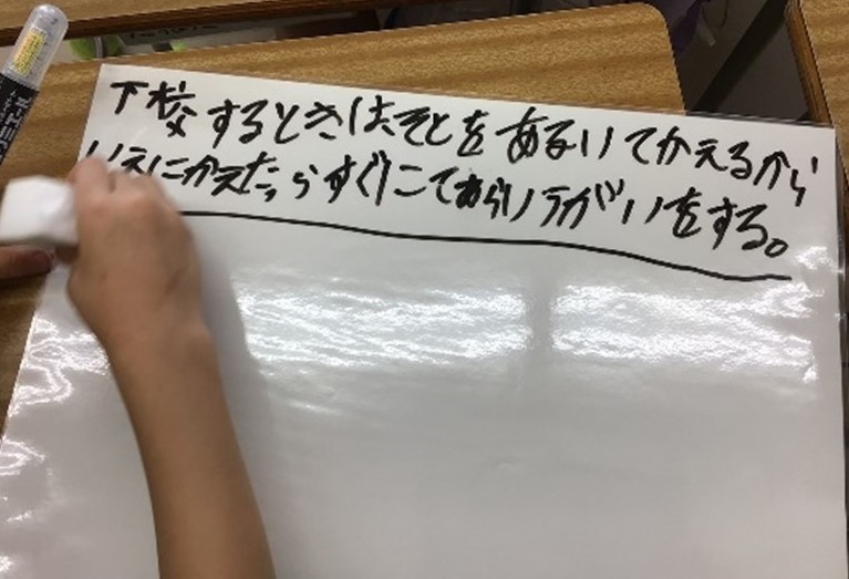 グループでの話合い。ミニホワイトボードに、「下校をするときは、外を歩いて帰るから、家に帰ったらすぐに手洗い・うがいをする。」と書いている。